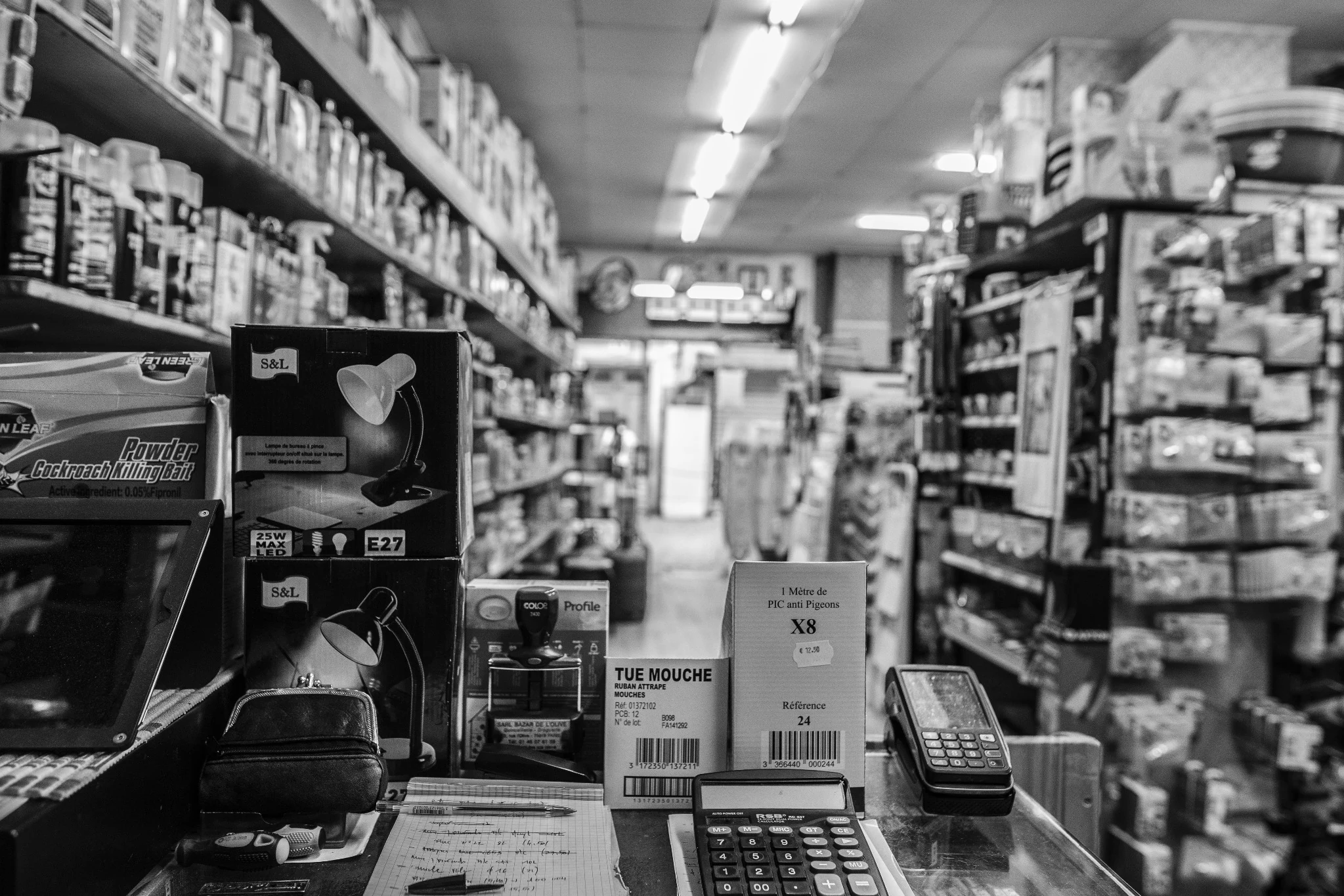 Un commerçant dans une quincaillerie remplie d’objets, en noir et blanc.